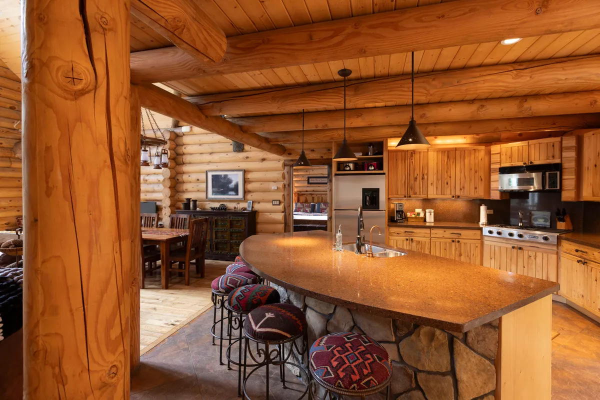 This image showcases a large kitchen island surrounded by wooden beams and stone accents. Bar stools with colorful fabric invite casual dining or socializing, and the open layout connects the kitchen with adjacent living spaces. The mood is warm and welcoming, perfect for gatherings and culinary adventures.