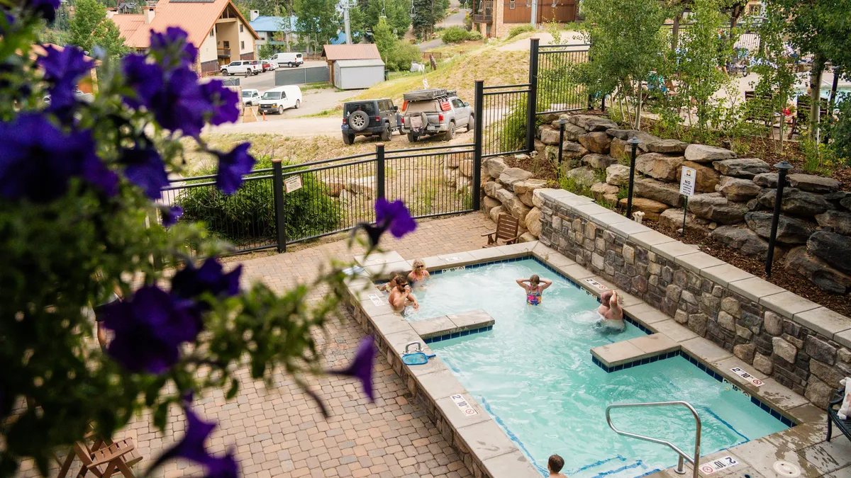 A lively outdoor resort scene featuring a swimming pool and hot tub surrounded by greenery and flowering plants. Children are seen playing in the pool, while adults relax in the hot tub and socialize nearby. The area is bathed in sunlight, conveying an atmosphere of joyful recreation and relaxation.