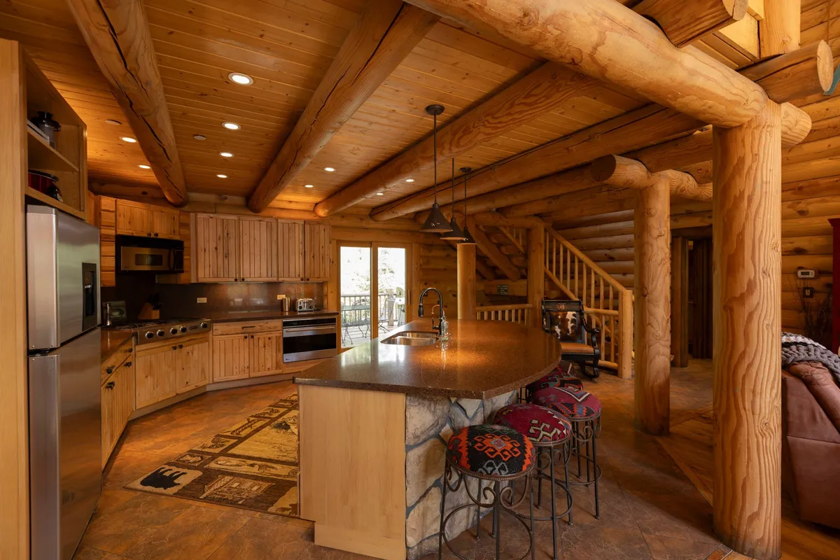 A close-up view of the kitchen features log beams, a central island with a stone countertop, and gleaming stainless steel appliances. The space feels cozy and rustic, inviting guests to prepare and enjoy meals together.