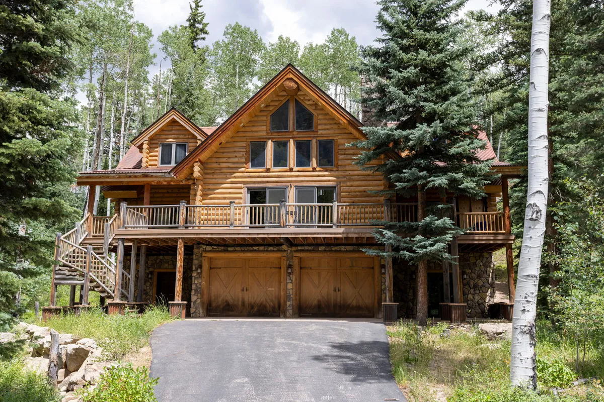 The hero image shows a charming log cabin-style townhome with a spacious front porch, stone-accented garage doors, and tall pine trees surrounding the property. The golden glow of sunset bathes the scene, inviting guests to imagine relaxing outdoors in tranquil mountain surroundings.