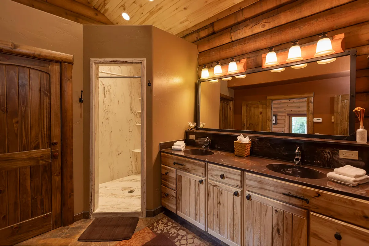 A spacious bathroom with a double-sink vanity, large mirror, shower area, and rustic wooden door, all bathed in warm, inviting lighting. The layout ensures ease and comfort for group or family use.