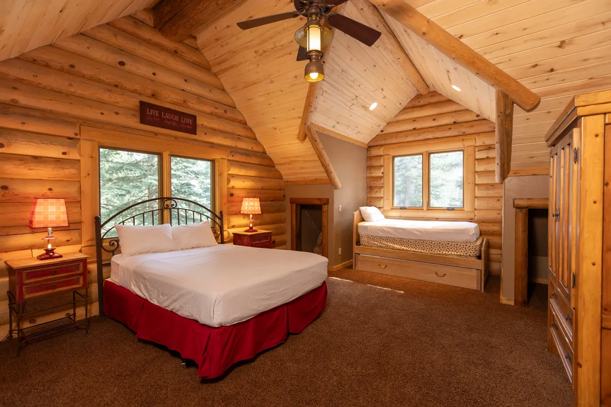 A family-friendly bedroom with wooden log walls, a queen bed with a red skirt, a bunk bed, bedside lamps, and large windows letting in natural light. The arrangement is both practical and welcoming, ideal for accommodating adults and children together.