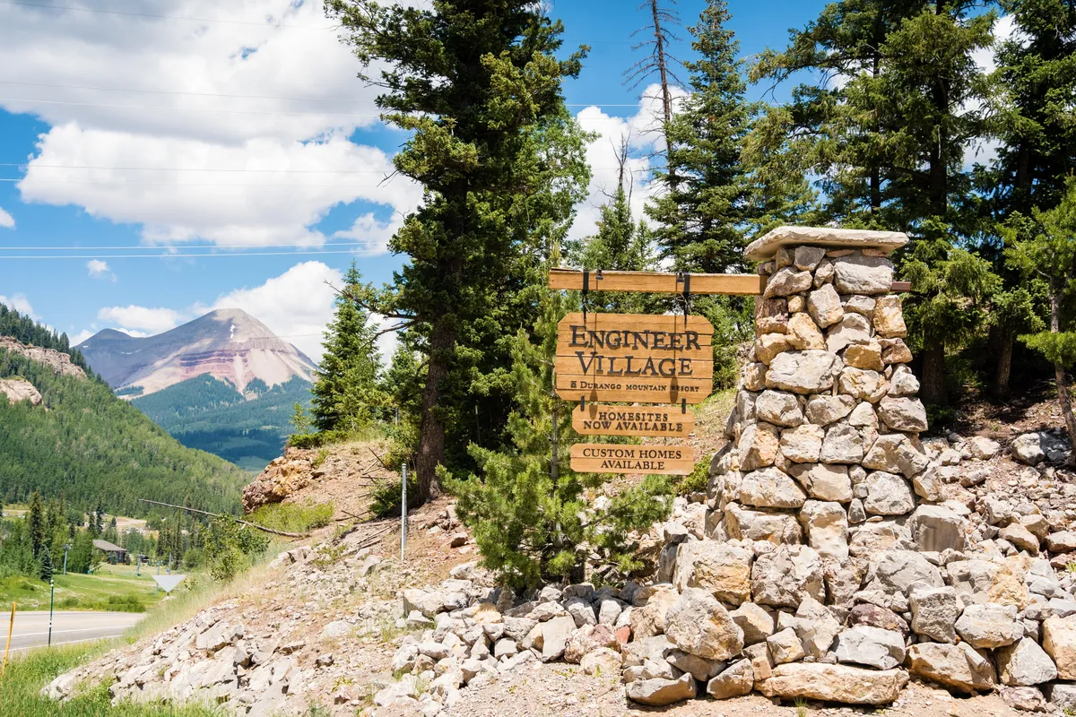 The exterior view showcases a sign for Engineer Village, with a backdrop of majestic mountains and a tree-lined setting. The scene exudes an adventurous and welcoming mood, suggesting easy access to nature, trails, and the broader resort community.