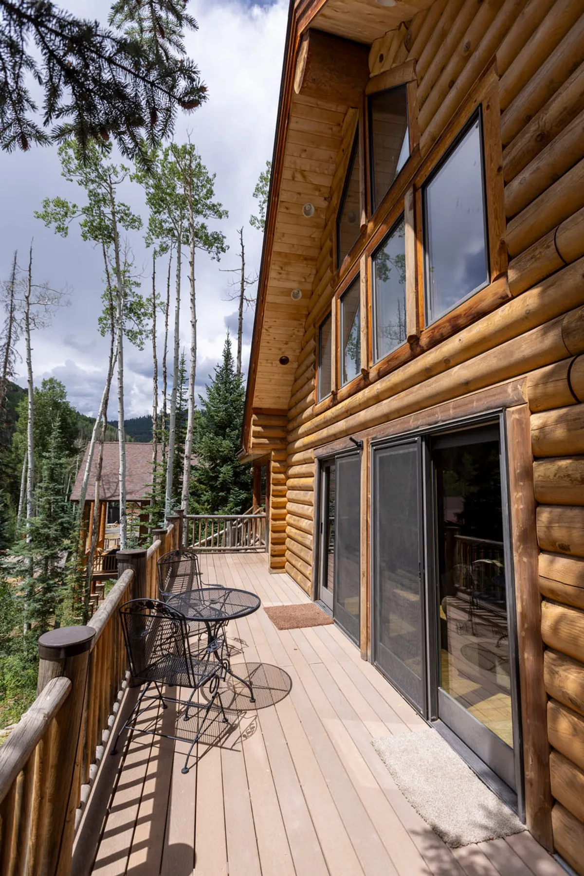 This exterior image captures the log cabin structure with an inviting outdoor seating area, set among beautiful trees and overlooking the mountain landscape. The setup suggests a peaceful environment for socializing or enjoying the view.
