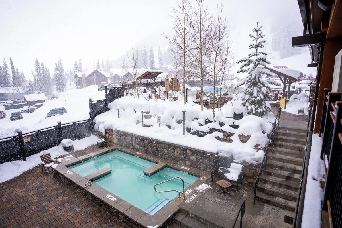 This image shows an outdoor hot tub set amid a snow-covered landscape, surrounded by trees and a wooden staircase leading down to the area. The scene exudes tranquility and the allure of resort amenities, inviting guests to relax after a day of adventure.