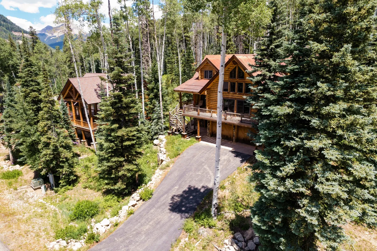 This exterior image features two log cabins nestled among aspen trees with a stunning mountain backdrop. The scene captures a peaceful natural setting ideal for outdoor activities, with visible hiking paths inviting exploration and opportunities for family gatherings or nature walks.