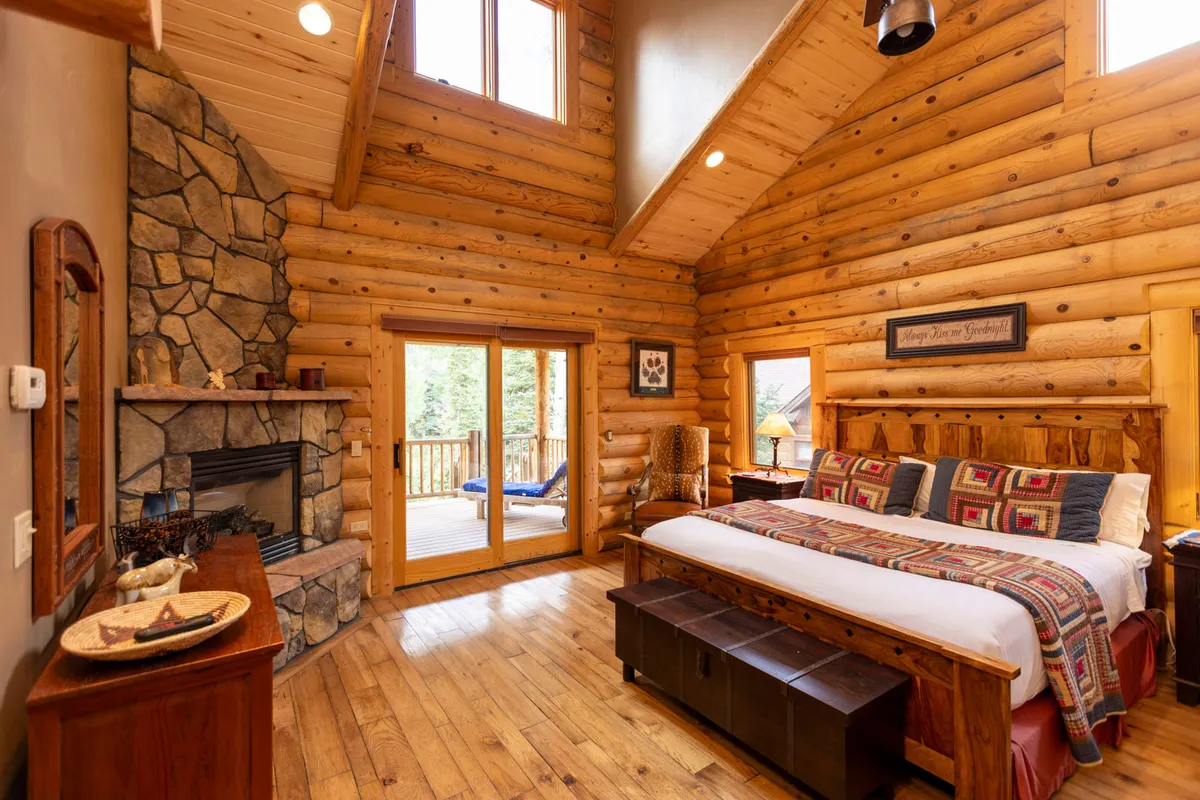 This inviting master bedroom features a large wooden bed, a stone fireplace, sliding glass doors leading to a deck, and decorative wall art, all set against a backdrop of warm wood tones and natural textures. The space exudes comfort and provides a restful retreat, with ample natural light streaming in.