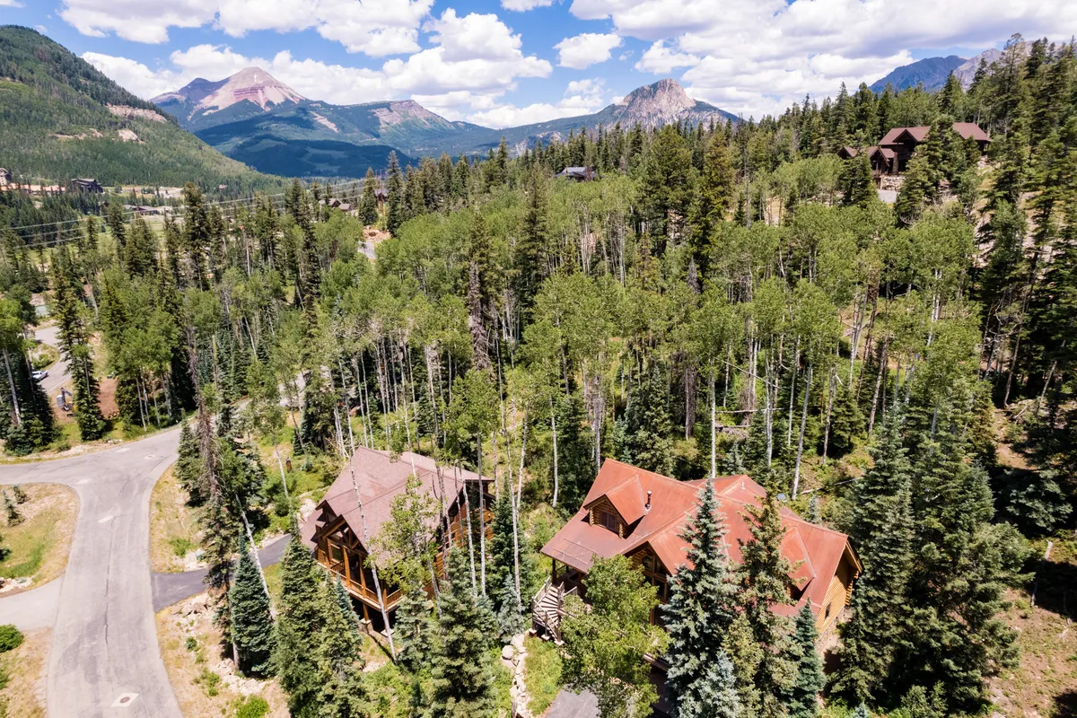 An exterior perspective showing two distinct houses nestled among trees with a mountain backdrop, encapsulating the tranquil surroundings and offering a sense of place. The open outdoor area hints at opportunities for family exploration and group activities in nature.