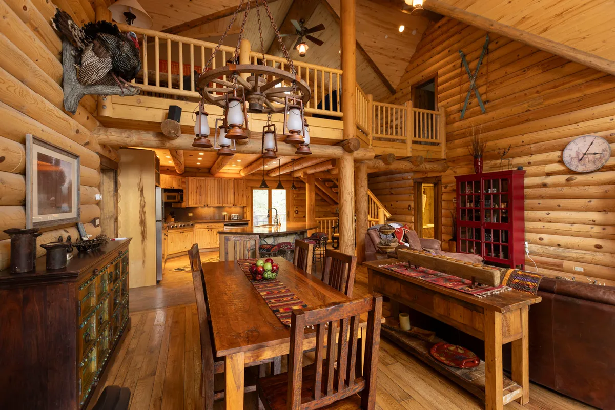 A large wooden dining table sits beneath an artful chandelier with lanterns. The table is set within a log-constructed dining area, with red accent furniture providing bursts of color. The warm, spacious environment is ideal for family gatherings, shared meals, and celebrations.
