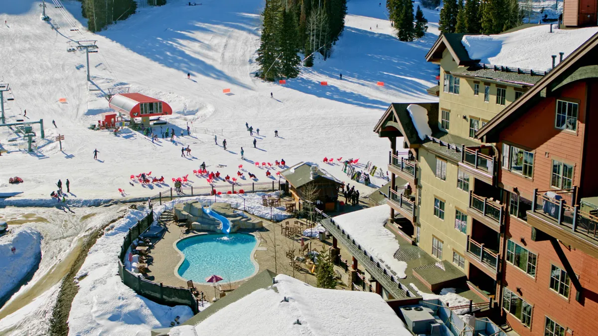 An action-filled resort environment with snow-covered ski slopes, a pool area, ski lifts, and striking building architecture. People are seen enjoying winter activities such as skiing and snowboarding, with the pool area in the foreground suggesting après-ski relaxation.